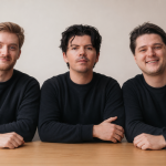 Three caucasian men in black long-sleeved crewneck shirts sit at a desk against a beige background.